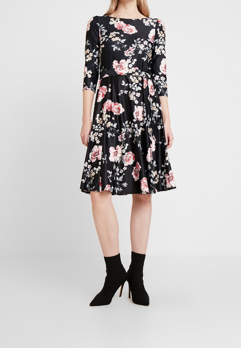 Femme portant une robe noire mi-longue avec imprimé floral rose et blanc et manches trois-quarts, associée à des bottines noires à talons hauts.