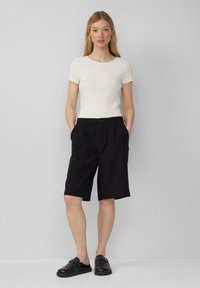 Weißes geripptes Kurzarmoberteil und schwarze knielange Shorts mit zwei Seiten Taschen, flacher Vorderseite und dezenten Nähdetails. Schwarze Slipschuhe.