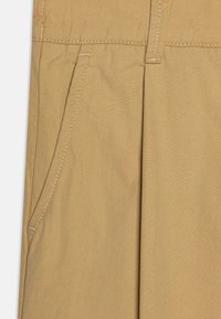 Pantaloni beige con una texture liscia, dotati di una tasca laterale e dettaglio con doppia piega. I punti di cucitura sono di un colore chiaro in contrasto.