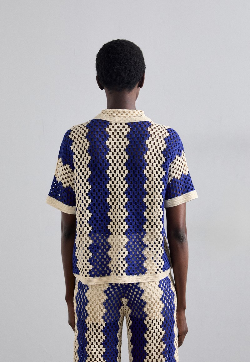Häkelshirt in Creme und Dunkelblau, mit einem karierten Muster, Polokragen und kurzen Ärmeln, getragen mit passenden Hosen.