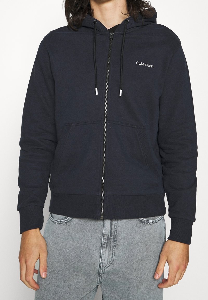 Sudadera con cremallera azul marino, con un bolsillo tipo canguro en la parte frontal, capucha con cordón, puños acanalados y logo bordado en el lado izquierdo del pecho. Fabricada en mezcla de algodón.