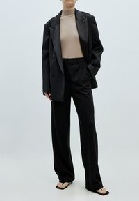 Zwarte oversized blazer met opvallende revers, een beige hoge nek top en losse zwarte broek. Draag het met zwarte flip-flop sandalen.