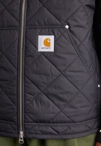 Sort quiltet jakke med en sølv lynlås, label med orange logo og trykknapper. Den har en tekstureret finish og struktureret design.