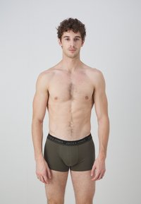Olivgrüne Boxer-Shorts mit einem schwarzen Bund, der den Schriftzug "JACK & JONES" trägt. Glatte Textur, taillierte Passform. Das Modell steht aufrecht.