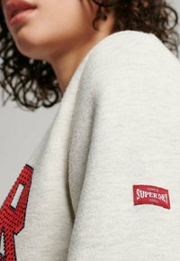 Zbliżenie na osobę w jasnoszarym swetrze z czerwoną naszywką "SUPERDRY" na rękawie i dużą czerwoną literą z przodu.