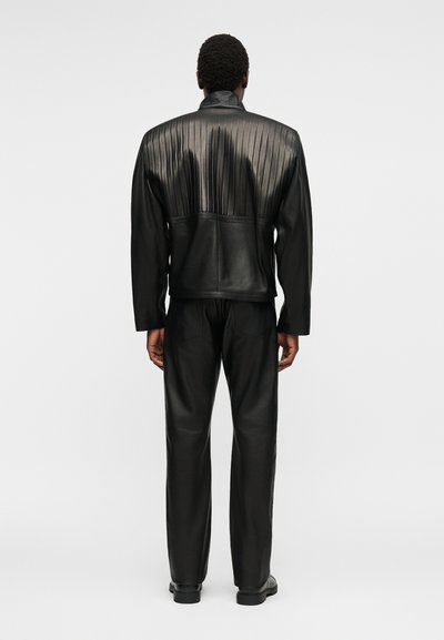 Άνδρας που φοράει μαύρο δερμάτινο τζάκετ με πιέτες και αντίστοιχο δερμάτινο παντελόνι, στέκεται πλάτη σε απλό φωτεινό φόντο.