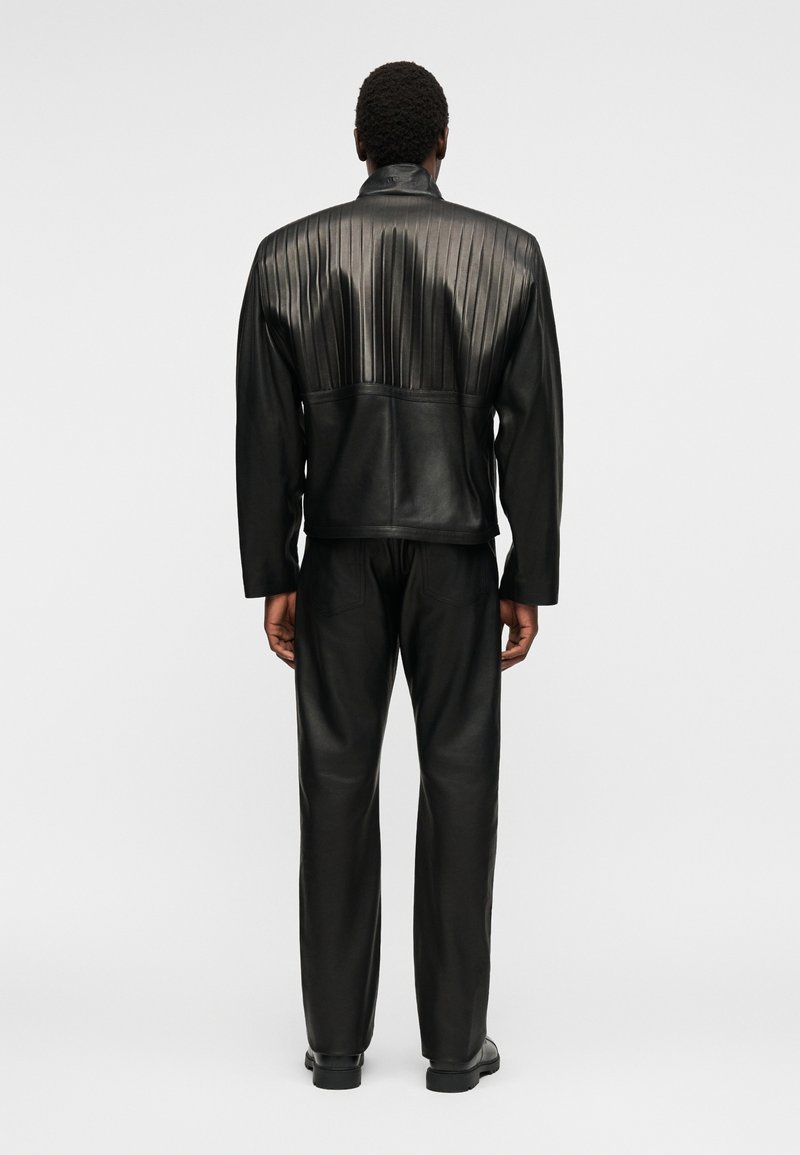 Homme portant une veste en cuir plissée noire et un pantalon en cuir assorti, debout dos à un fond clair uni.