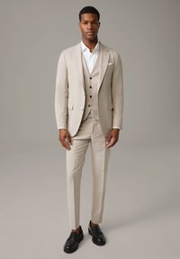 Beige Dreiteiler mit strukturiertem Finish, bestehend aus einem taillierten Sakko, einer Weste mit dunklen Knöpfen und schmalen Hosen. Kombiniert mit schwarzen Slippern.