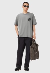 Camiseta gris de gran tamaño con un diseño gráfico negro, pantalones cargo negros y zapatillas negras. Lleva una bolsa acolchada de color verde oliva.