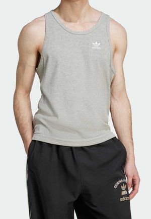 Mann trägt ein hellgraues Adidas-Top und schwarze Adidas-Trainingshose, steht mit den Händen in den Taschen vor einem schlichten hellen Hintergrund.