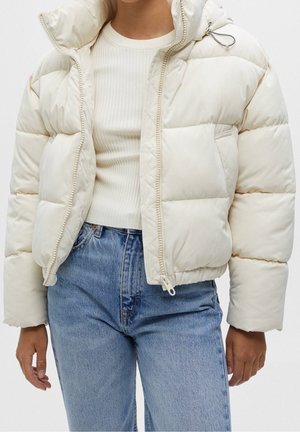 Person wearing a cream-colored puffer jacket over a white ribbed sweater and light blue high-waisted jeans, standing with arms relaxed.