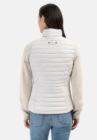 Weiße, gesteppte Weste mit hohem Kragen, mit Druckknopfakzenten auf der Rückseite, kombiniert mit einem hellen, strukturierten Pullover und blauen Jeans.