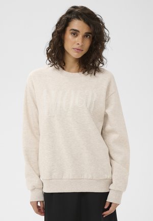 Kvinde med krøllet hår iført en beige sweatshirt med perlebesat "AMOUR" tekst og en sort nederdel mod en ensartet lys baggrund.