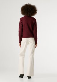 Μπορντό πλεκτό πουλόβερ με υφασμάτινα μανίκια, συνδυασμένο με λευκά παντελόνια με φαρδιά γραμμή και μαύρες γόβες, απεικονιζόμενο από πίσω.