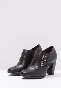 Botas de tobillo de cuero negro con punta curva, dos correas con hebillas y adornos de piedras brillantes en la punta. Tacón grueso y alto.