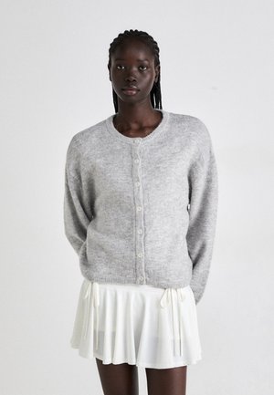 Grijze gebreide cardigan met een ronde halslijn, knopen aan de voorkant en lange mouwen, gedragen over een witte uitlopende rok. Zachte textuur en casual ontwerp.