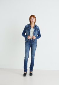 Veste en denim avec une coupe classique, de couleur bleu clair, et deux poches avant, associée à un jean ajusté noir et des chaussures noires.