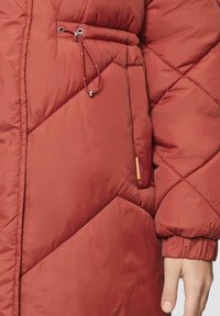 Rode gewatteerde pufferjas met een trekkoord in de taille, elastische manchetten en een zichtbaar merketiket. Het materiaal lijkt glad met een matte afwerking.