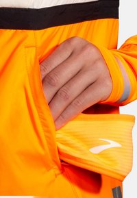Bright orange jacket with black accents and a reflective stripe, featuring a hand reaching into a pocket. Textured, lightweight fabric visible.