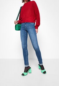 Donna che indossa un maglione rosso, jeans skinny blu, sandali alti verdi con calze nere in pizzo, e porta una borsa a tracolla verde trapuntata.