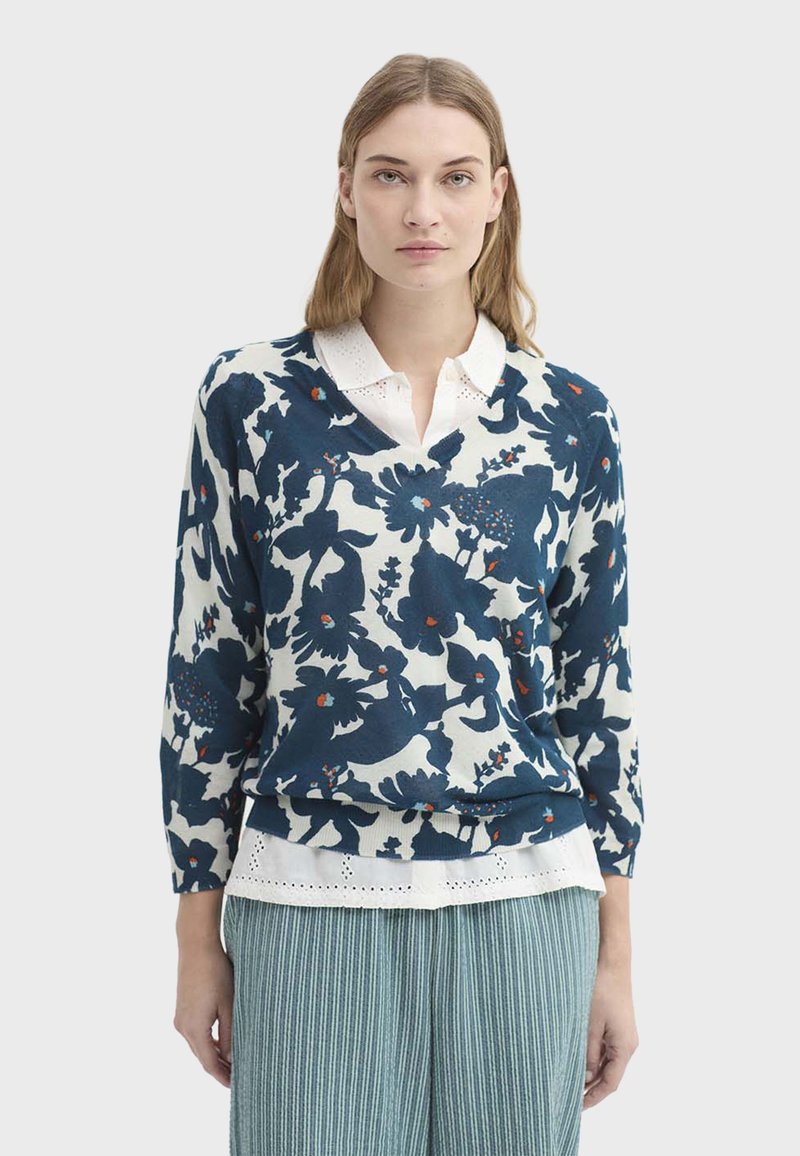 Femme portant un chemisier blanc sous un pull bleu marine à motif floral avec des manches trois-quarts et un pantalon bleu clair rayé.