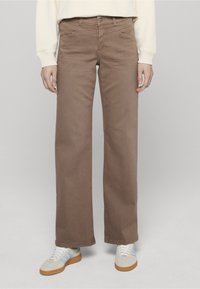 Brązowe spodnie o szerokich nogawkach z gładką teksturą, pięcioma kieszeniami i zapięciem na guzik. W połączeniu z białymi sneakersami z niebieskimi akcentami.