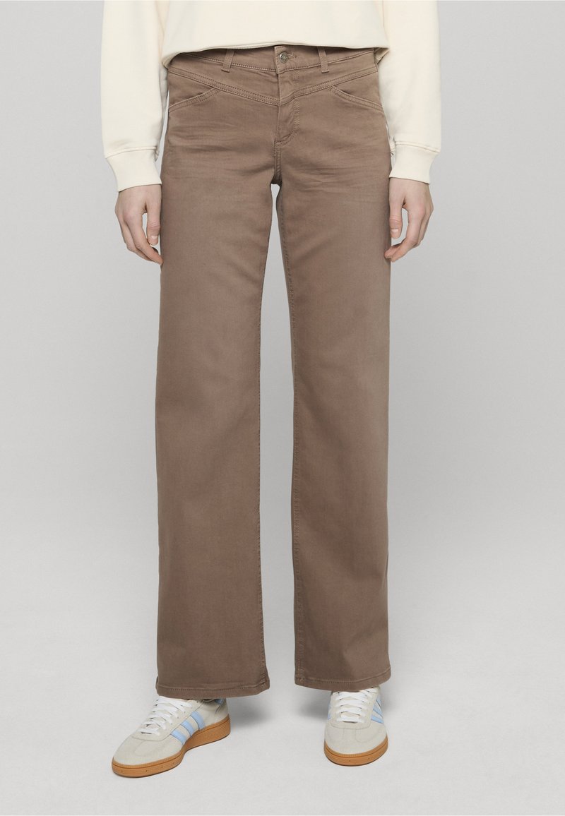 Brązowe spodnie o szerokich nogawkach z gładką teksturą, pięcioma kieszeniami i zapięciem na guzik. W połączeniu z białymi sneakersami z niebieskimi akcentami.