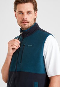 Man with beard wearing a teal and navy fleece vest over a white T-shirt, holding the zipper near his chin.