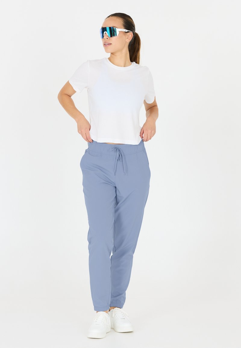 Witte cropped t-shirt, lichtblauwe elastische broek met zakken en witte sneakers. Model draagt blauw-getinte zonnebril.
