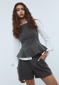 Sivi prugasti peplum top uparen s odgovarajućim šortsevima. Karakterizira ga strukturirani dizajn, glatka tekstura i slojeviti rubni detalj. Ispod je bijela košulja.