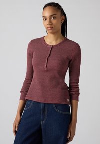 Camicia henley a maniche lunghe di colore bordeaux con un motivo testurizzato, scollatura rotonda, bottoni sul davanti e vita aderente. Abbinata a jeans in denim scuro.