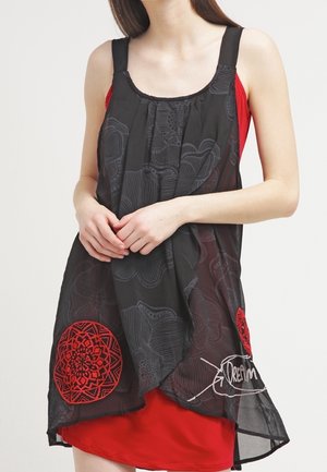 Mujer vistiendo un vestido rojo sin mangas con una capa negra translúcida y suelta que presenta patrones circulares rojos y la palabra "Dream" en la parte inferior derecha.