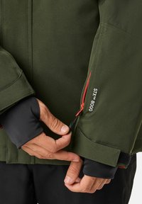 Grüne wasserdichte Jacke mit schwarzen Bündchen, ausgestattet mit einer Reißverschlusstasche mit roten Akzenten und verstellbaren Riemen zur Verbesserung der Passform.