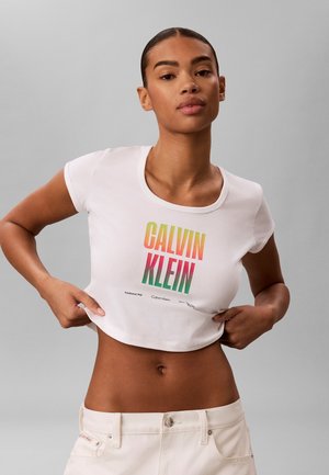 Kobieta podnosi białą krótka koszulkę z tęczowym logo "Calvin Klein", nosi jasne spodnie, stoi na jednolitym szarym tle.