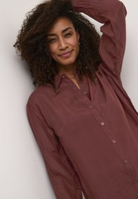 Camicia button-up color bordeaux realizzata in tessuto liscio, con maniche lunghe e vestibilità rilassata. Include un colletto e dettagli in bottone sulla parte anteriore.