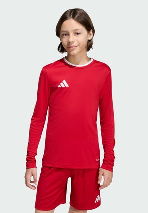 Enfant portant un maillot de sport rouge à manches longues et un short assorti avec des logos Adidas blancs sur un fond uni.