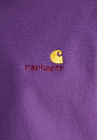 Lilla bomuldsstof med glat tekstur, der har et broderet gult og burgunder Carhartt-logo. Designet er enkelt og dristigt.