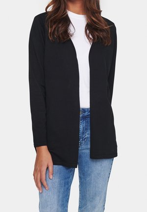 Kobieta ubrana w czarny otwarty sweter na biały top i niebieskie dżinsy, stojąca na tle jasnego jednolitego tła.