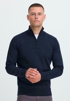 Pull en maille bleu marine avec un col haut et une fermeture éclair quart, présentant des manches longues et une coupe ajustée. Texture douce avec une étiquette sur le côté.