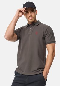 Polo de color gris oscuro con un collar clásico, mangas acanaladas, detalles rojos y un pequeño logo bordado en el pecho. Combinado con pantalones cortos azul marino.