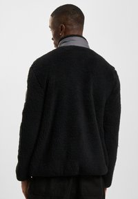 Chaqueta de forro polar negra con cuello gris, con un exterior texturizado y un interior suave. Corte holgado con mangas largas y sin cierres.