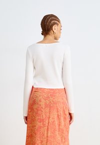 White long-sleeve top with a textured pattern, paired with a floral-patterned skirt in shades of orange and pink, featuring a back button detail.