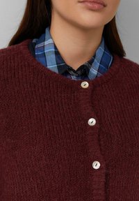 Burgunderfarbener Strickpullover mit strukturiertem Erscheinungsbild, rundem Halsausschnitt und drei weißen Knöpfen vorne, über einem blauen Karoshemel getragen.