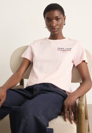 T-shirt en coton rose clair avec des manches courtes et un col rond, affichant le logo "GANT 1949" en bleu marine et rouge sur le devant. Coupe décontractée.