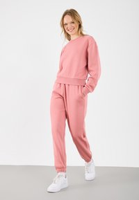 Hanorac din bumbac roz și pantaloni jogger asortați cu manșete cu nervuri, având buzunare laterale. Modelul poartă teneși albi. Fundal minimalist.