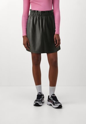 Persoon die een zwarte leren rok, een roze top met lange mouwen, witte sportsokken en zwart-witte hardloopschoenen draagt, staand op een witte vloer.