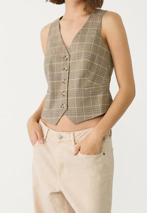 Vrouw draagt een mouwloos bruin geruit vest met knopen en lichtbeige broek met handen in de zakken, tegen een effen lichte achtergrond.
