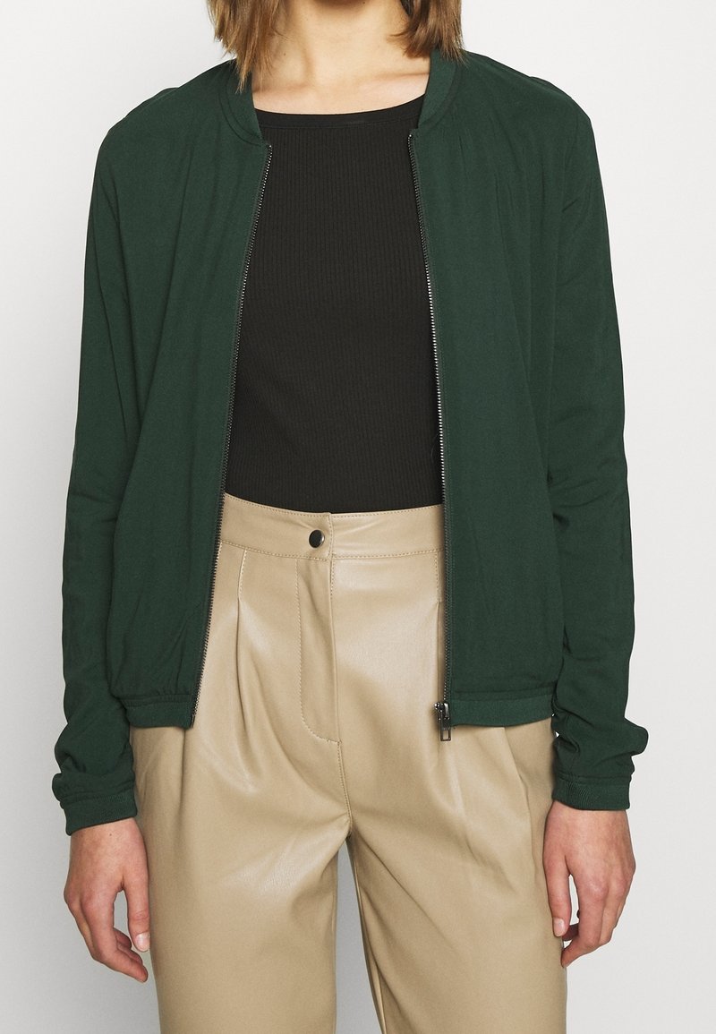 Veste bomber légère verte avec fermeture éclair frontale, poignets et col côtelés, associée à un pantalon beige taille haute en similicuir.