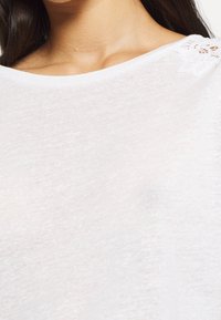 Bílý, lehký látkový top s kulatým výstřihem. Obsahuje jemnou květinovou krajkovou výzdobu na rameni. Měkčená textura, volný design.