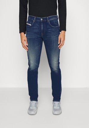 Jeans de mezclilla azul oscuro con un corte ajustado, que presentan un ligero desgastado, cinco bolsillos y un cierre de botón. Llevados con zapatillas deportivas grises.
